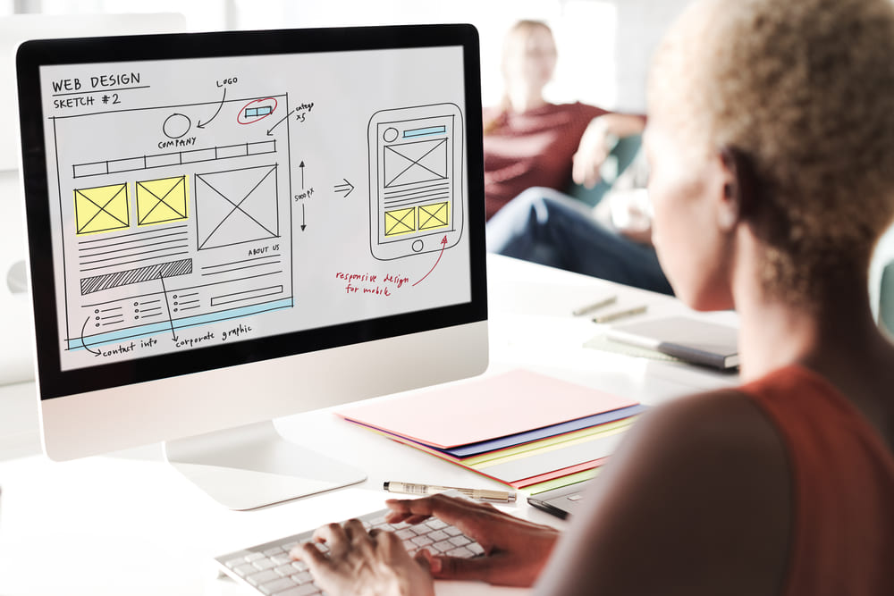 Mulher usando computador que está com um protótipo de layout e aplicativo na tela, simbolizando a diferença entre UX e UI - Design de interface