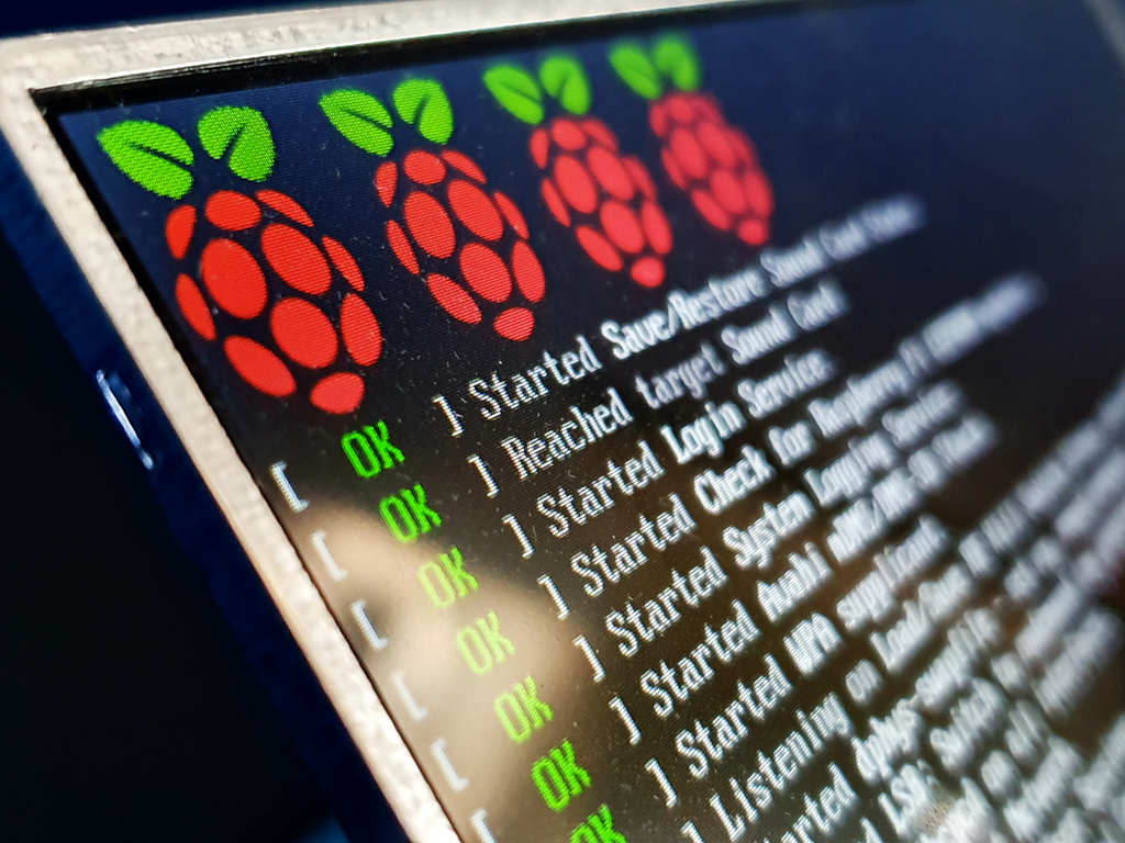 Display Raspberry-Pi mostrando "ok" para inicializações do sistema. Na parte superior da tela estão quatro logotipos, que são a ilustração de framboesas vermelhas