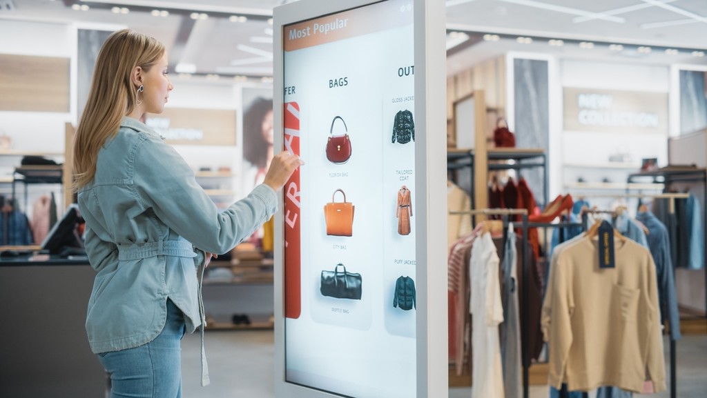 Dentro uma loja de roupas e acessórios, mulher usando display touch que exibe bolsas e roupas na tela, ilustrando um dos tipos de display