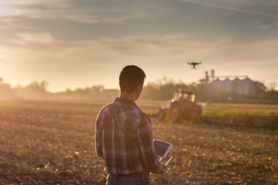 Olhando para plantação no horizonte, um trato e um drone, um homem de camisa xadrez está segurando um monitor de plantio (similar a um tablet branco, com duas antenas).