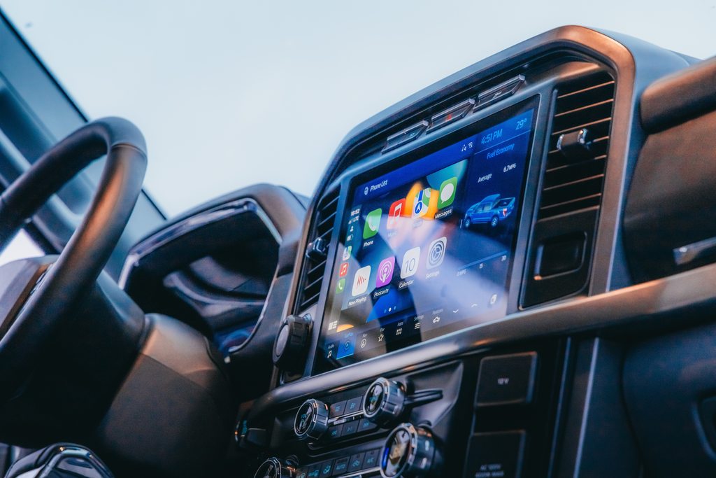 Interface multimidia no painel de um carro, a tela do monitor mostra o home com aplicativos como: Configurações, calendário, GPS, mensagens, música, podcasts e vídeo. Além de uma ilustração do carro com informações sobre ele.